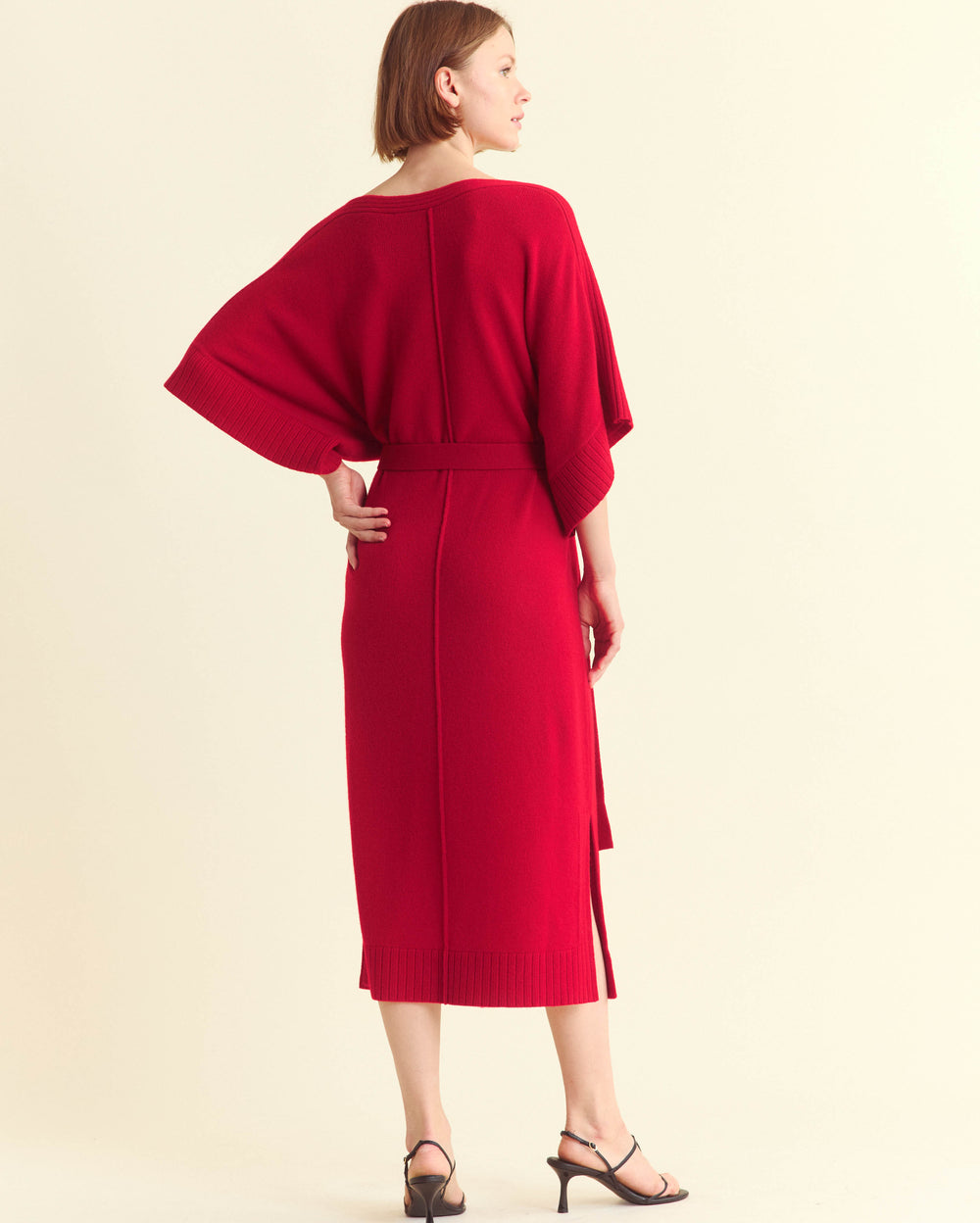 A woman stands facing away, wearing a mid-length, bright red knit dress with wide sleeves and a waist belt. She has short hair and is wearing black heeled sandals against a light background.