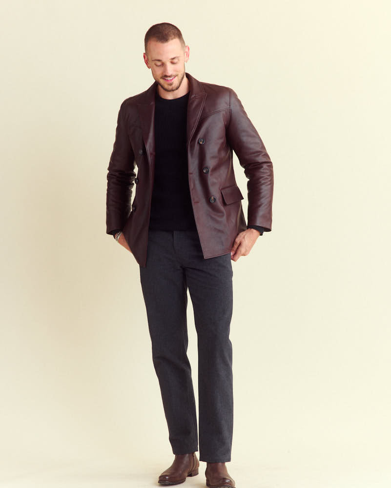 A man wearing a dark brown leather blazer, black sweater, charcoal gray pants, and brown shoes stands against a light beige background, looking down with a slight smile and one hand in his pocket.