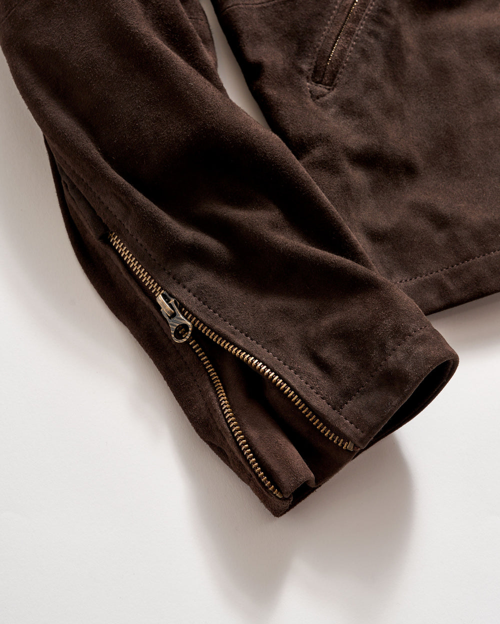 A close-up of a brown suede jacket sleeve with a visible gold zipper partially unzipped, lying on a white surface. The fabric appears soft and the stitching detail is visible.