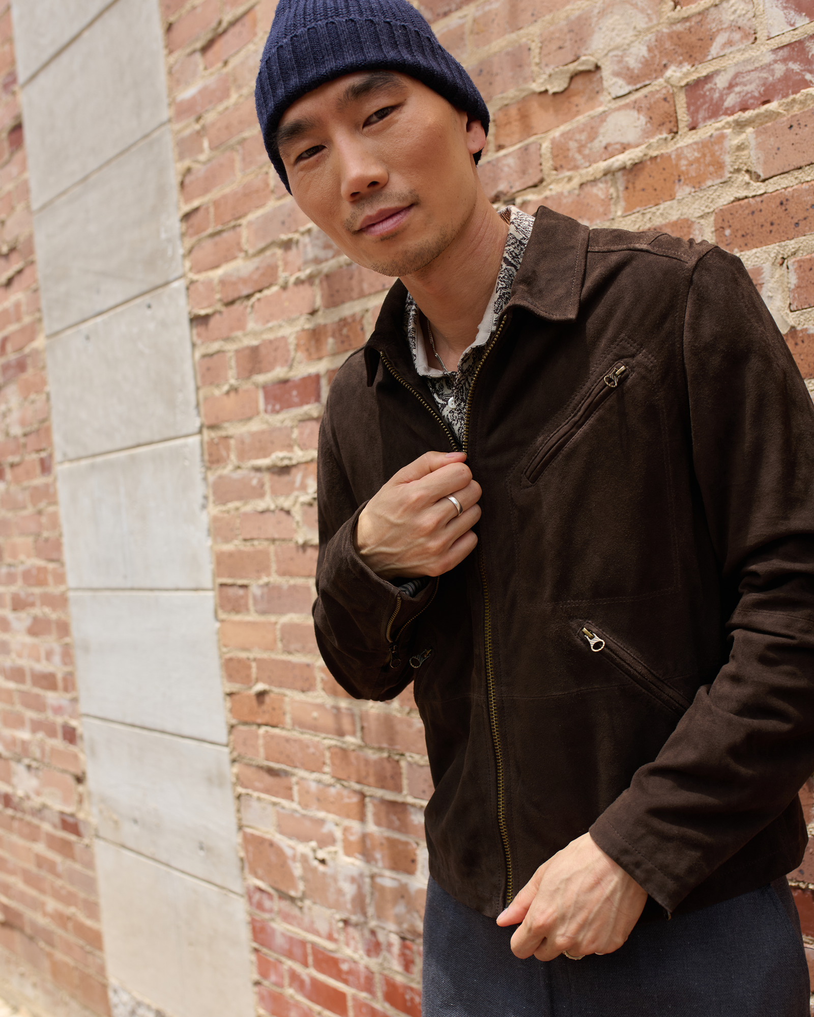 A person in a brown suede jacket and blue knit beanie stands in front of a brick wall, looking at the camera with a slight smile and holding the jacket’s lapel.