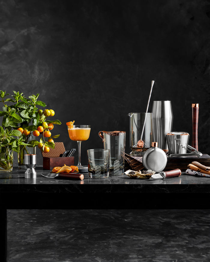 A black marble table set with cocktail-making tools, glasses, citrus fruits, a potted plant with oranges, a cocktail shaker, and a prepared orange cocktail, all against a dark background.
