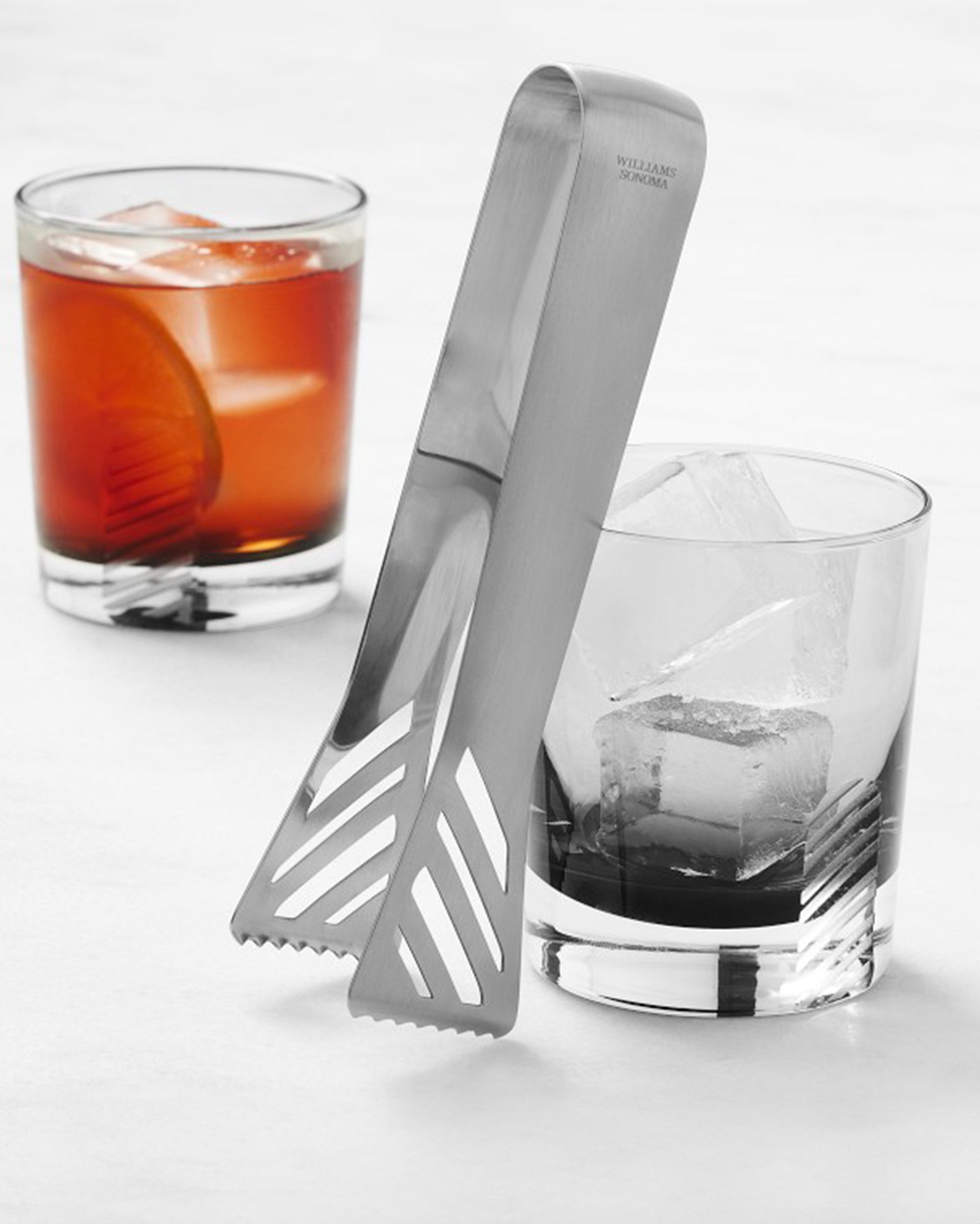Two glasses with drinks and ice cubes sit on a white surface; a pair of stainless steel tongs rests on one glass. One drink is clear, the other is reddish-brown with a citrus slice.