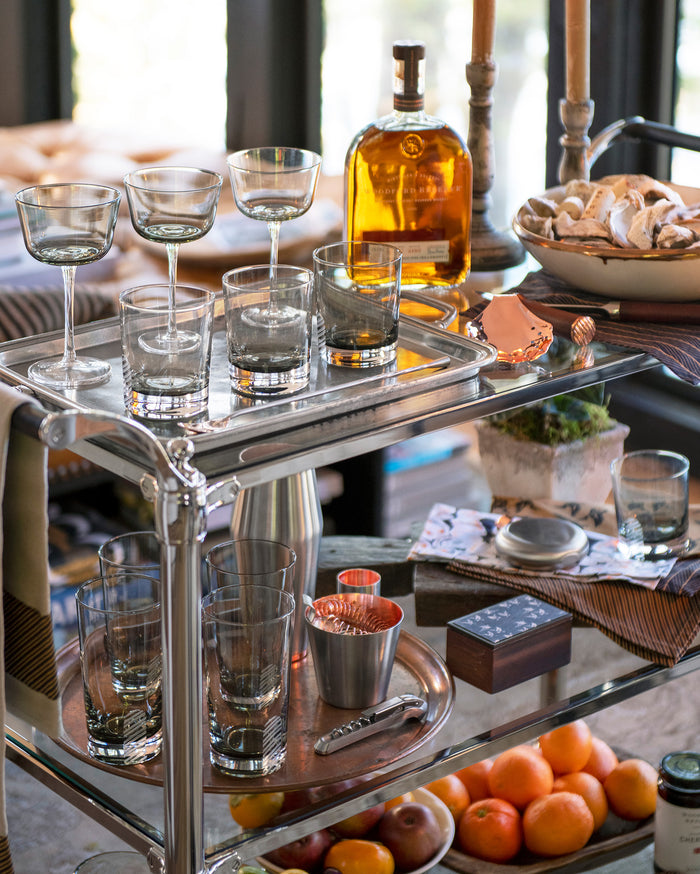 A silver bar cart with cocktail glasses, a bottle of whiskey, a bowl of cookies, a shaker, candles, a small potted plant, and a tray of fresh oranges on the lower shelf, set by a window.