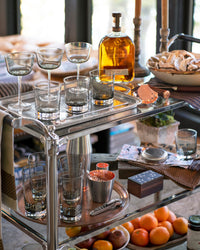 A bar cart holds various glasses, a bottle of liquor, a bowl of bread, a cocktail shaker, a copper cup, cocktail tools, and a small box, with a tray of oranges and other snacks on a table below.