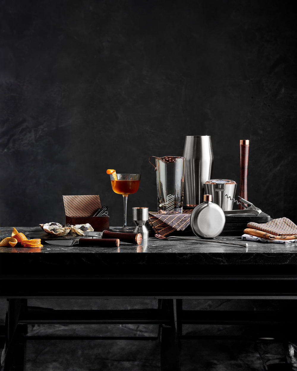 A collection of elegant barware, including a cocktail shaker, glass with an orange garnish, flasks, jigger, napkins, and oysters, arranged on a dark table against a dark, textured background.