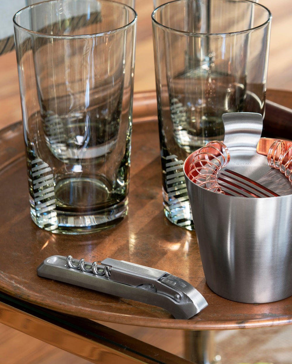 Two tall clear glasses, a metal cocktail strainer in a small stainless steel cup, and a metal corkscrew rest on a round copper tray with a wooden floor in the background.