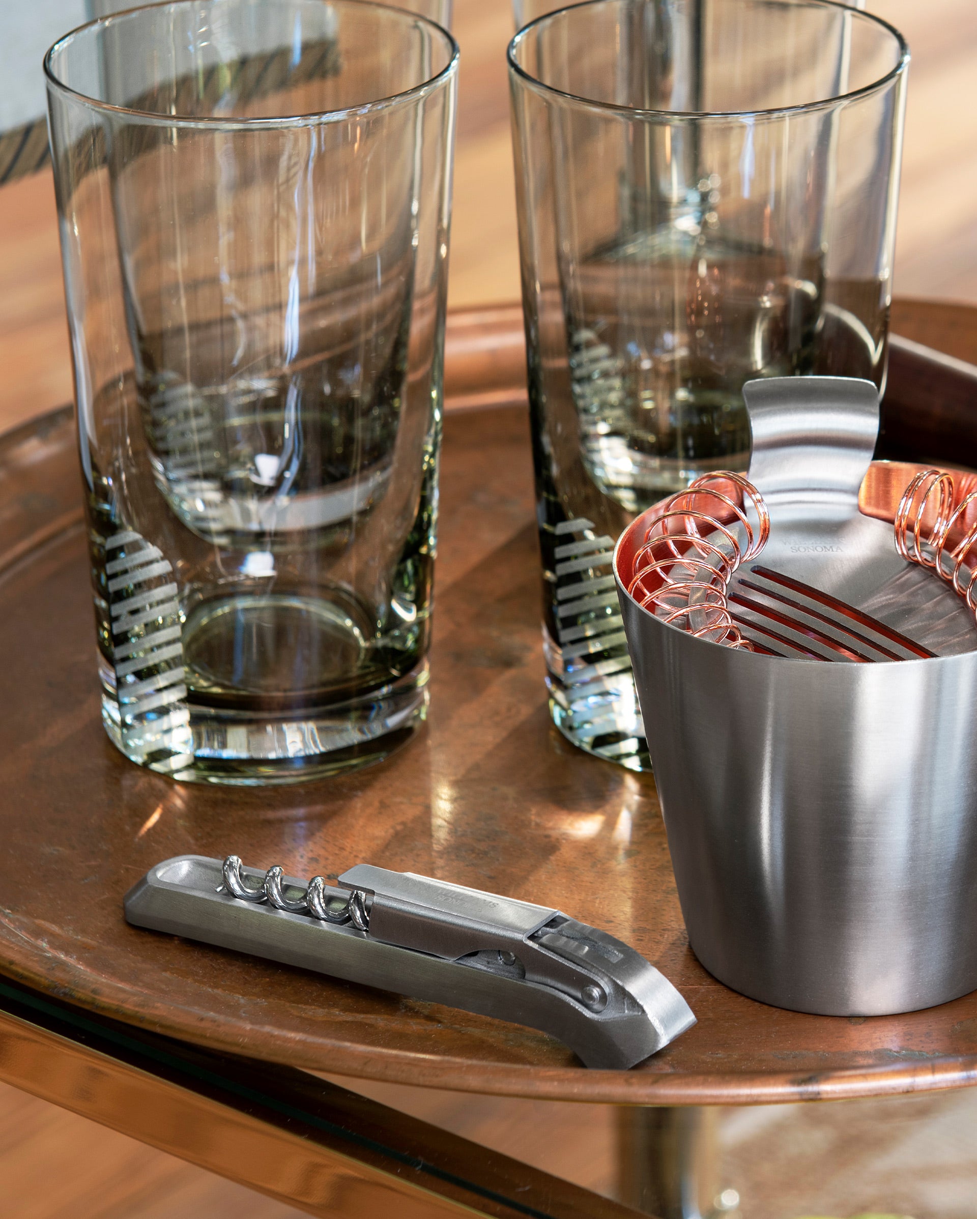 Two tall clear glasses, a metal cocktail strainer in a small stainless steel cup, and a metal corkscrew rest on a round copper tray with a wooden floor in the background.