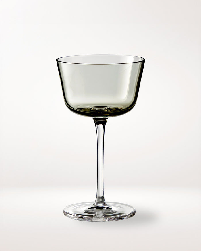 A single, elegant, clear wine glass with a wide bowl and long stem is centered on a plain, light background. The glass has a slightly smoky tint.