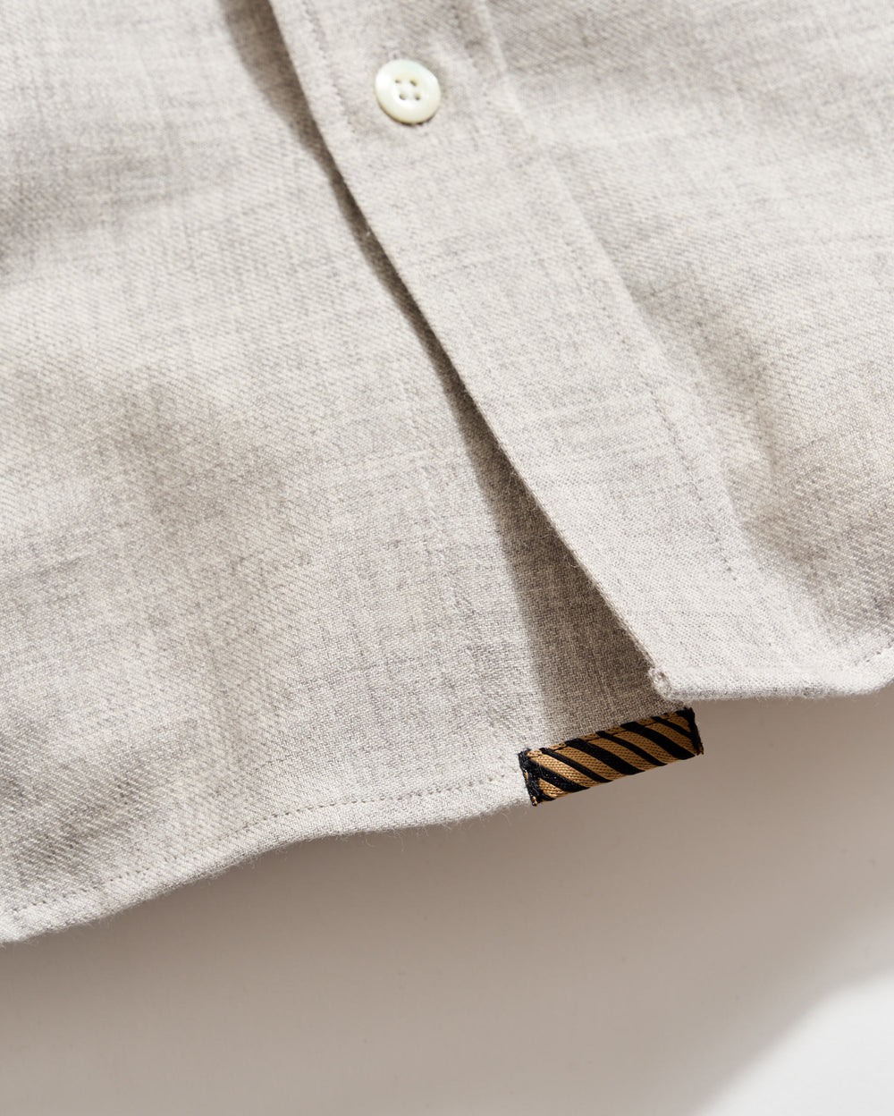 A close-up of a light beige shirt made of textured fabric, showing a white button and a small black tag with diagonal gold stripes near the hem.
