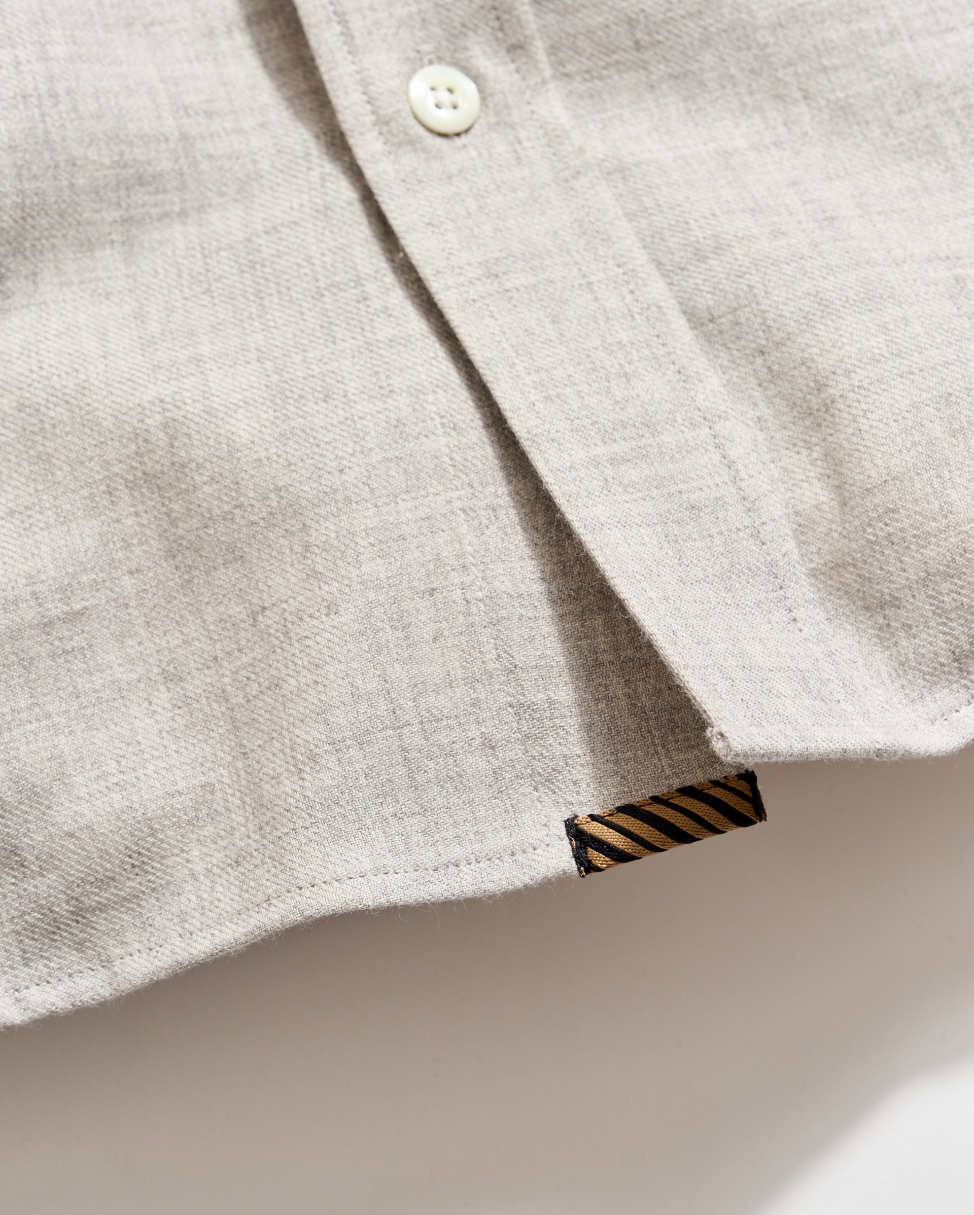 A close-up of a light beige shirt made of textured fabric, showing a white button and a small black tag with diagonal gold stripes near the hem.