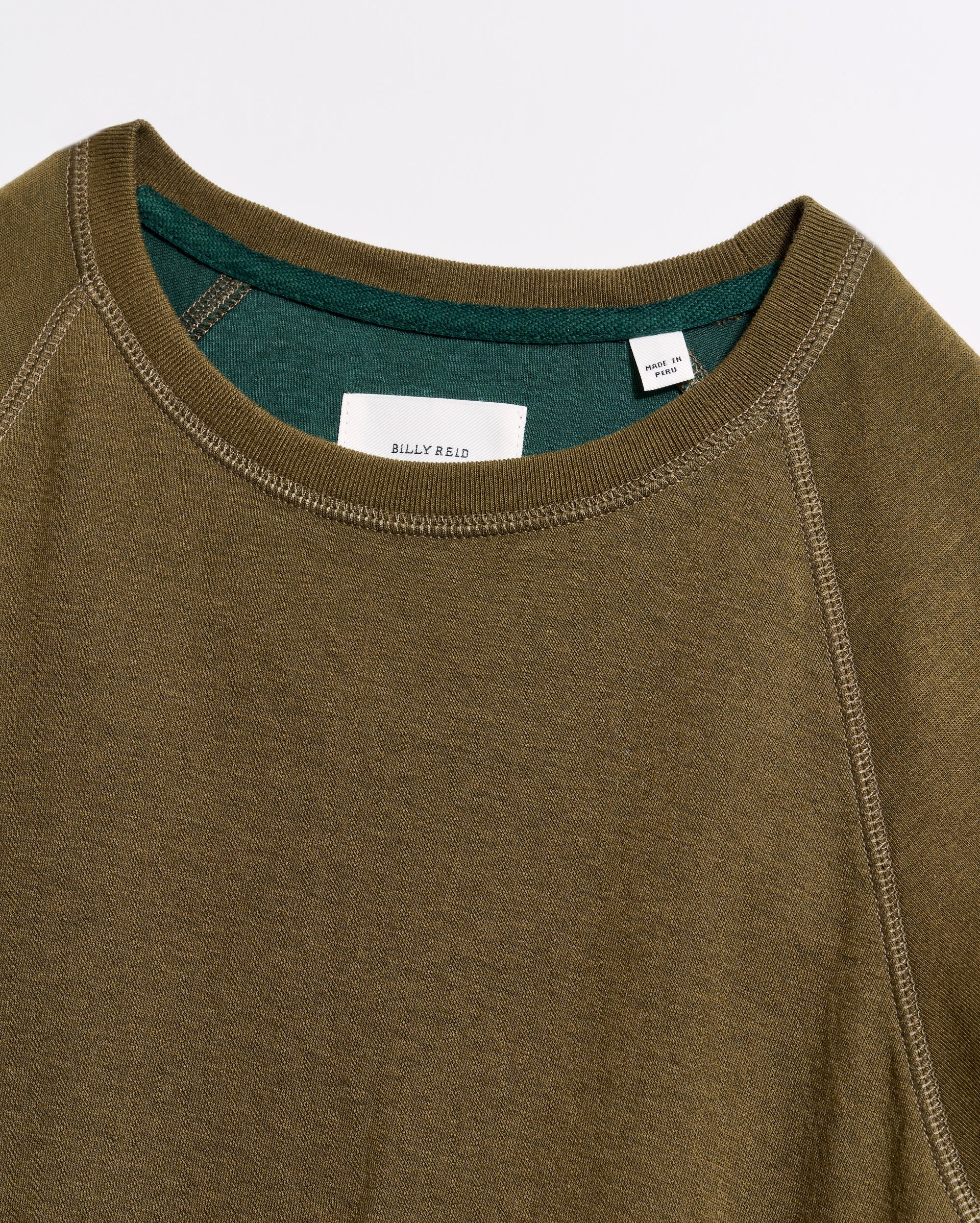 Close-up of an olive green crewneck sweatshirt with visible stitching and a contrasting green inner collar. A white tag with text is sewn inside the neckline. The fabric appears soft and casual.