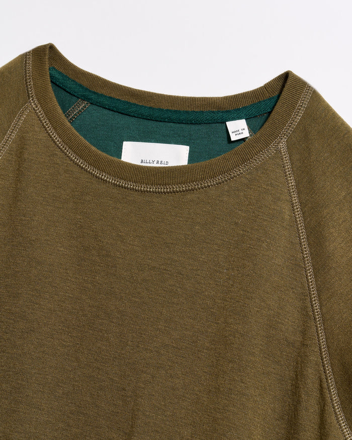 Close-up of an olive green crewneck sweatshirt with visible stitching and a contrasting green inner collar. A white tag with text is sewn inside the neckline. The fabric appears soft and casual.