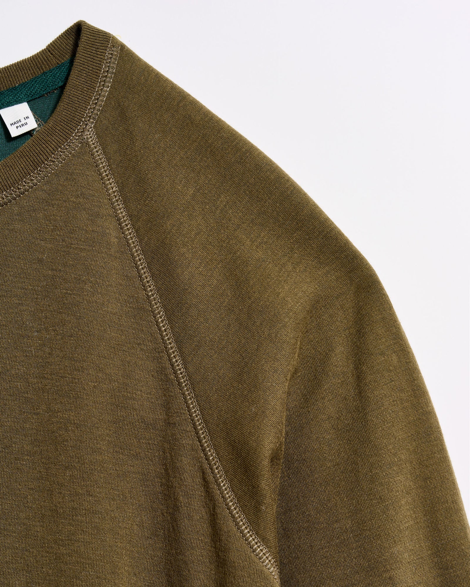 Close-up of an olive green sweatshirt made from textured fabric, showing the shoulder seam and part of the collar with a visible tag inside. The background is plain white.