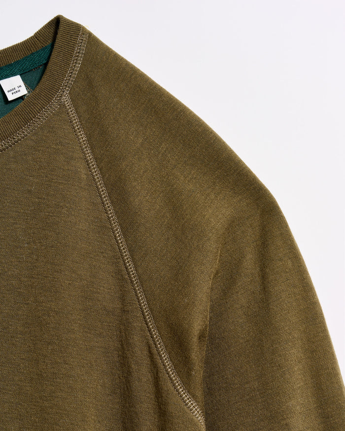Close-up of an olive green sweatshirt made from textured fabric, showing the shoulder seam and part of the collar with a visible tag inside. The background is plain white.