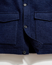 Close-up of a navy blue wool coat, showing two flap pockets, four black buttons, and part of the coats hem against a white background.