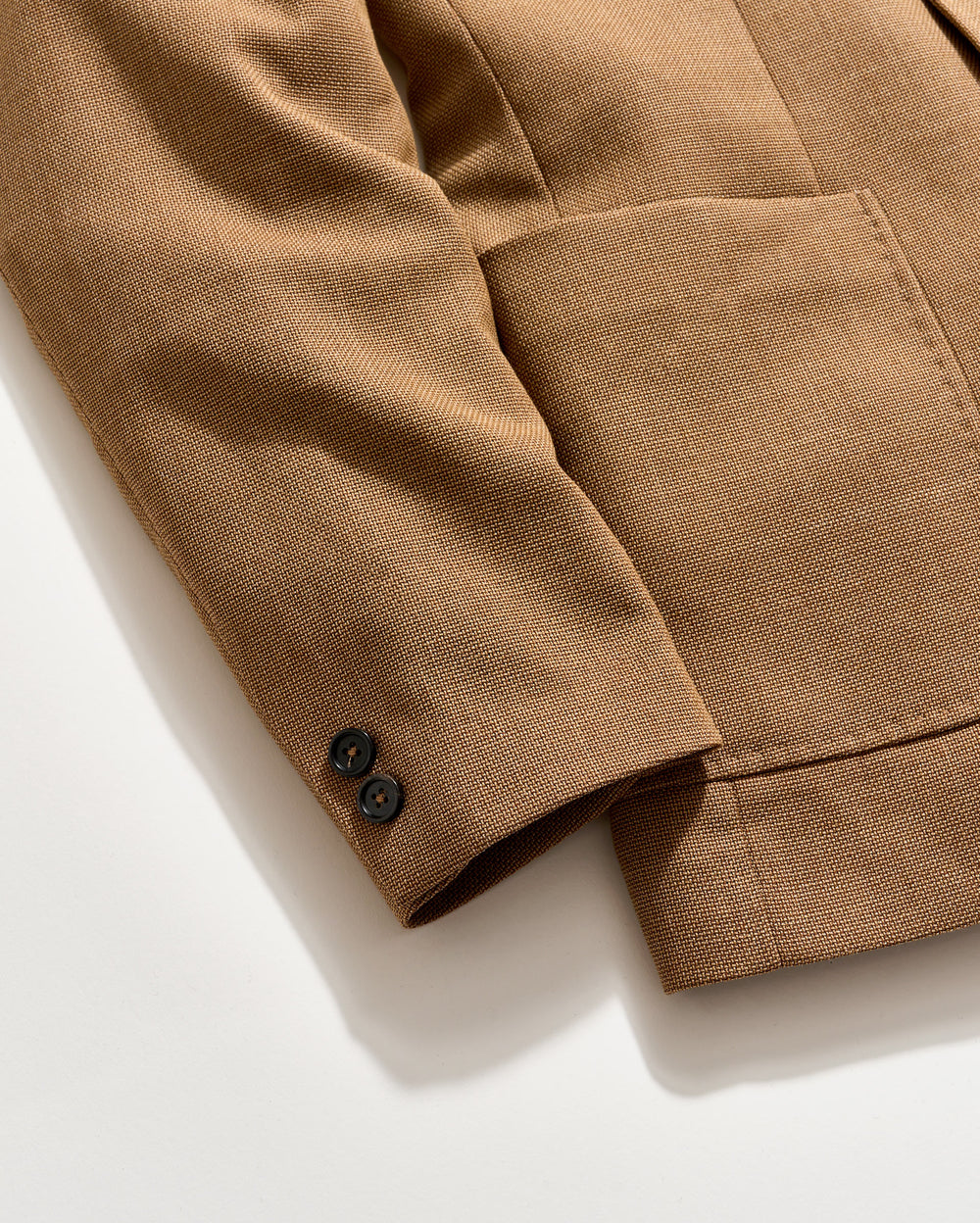 A close-up of a brown textured blazer sleeve with two black buttons on the cuff, shown on a light background. The blazer features a large front pocket.