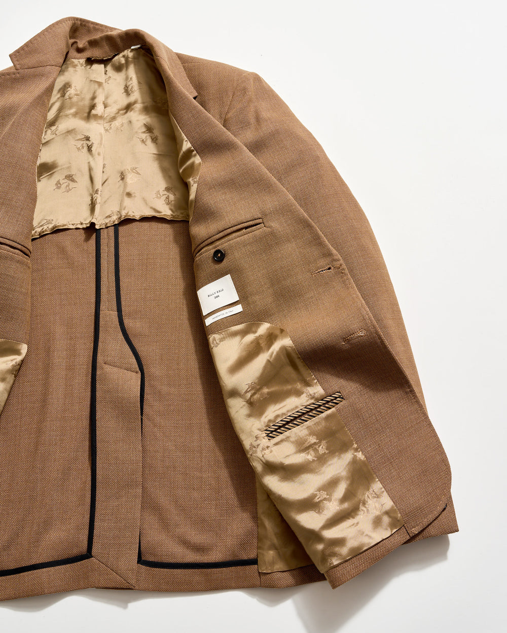 A brown blazer laid open on a white surface, showing its beige, patterned satin lining, multiple interior pockets, a button detail, and a white label sewn inside.
