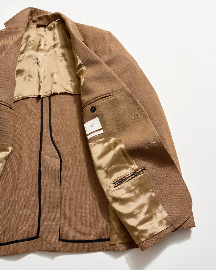 A brown blazer laid open on a white surface, showing its beige, patterned satin lining, multiple interior pockets, a button detail, and a white label sewn inside.