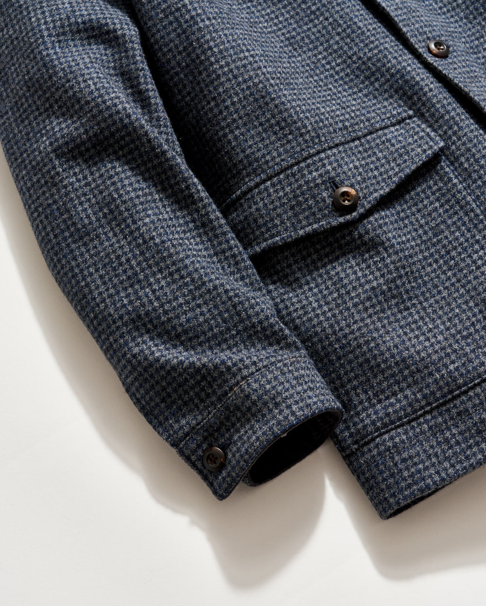 Close-up of a blue and gray houndstooth-patterned jacket, showing the sleeve, buttoned cuff, and a flap pocket with buttons on a white background.