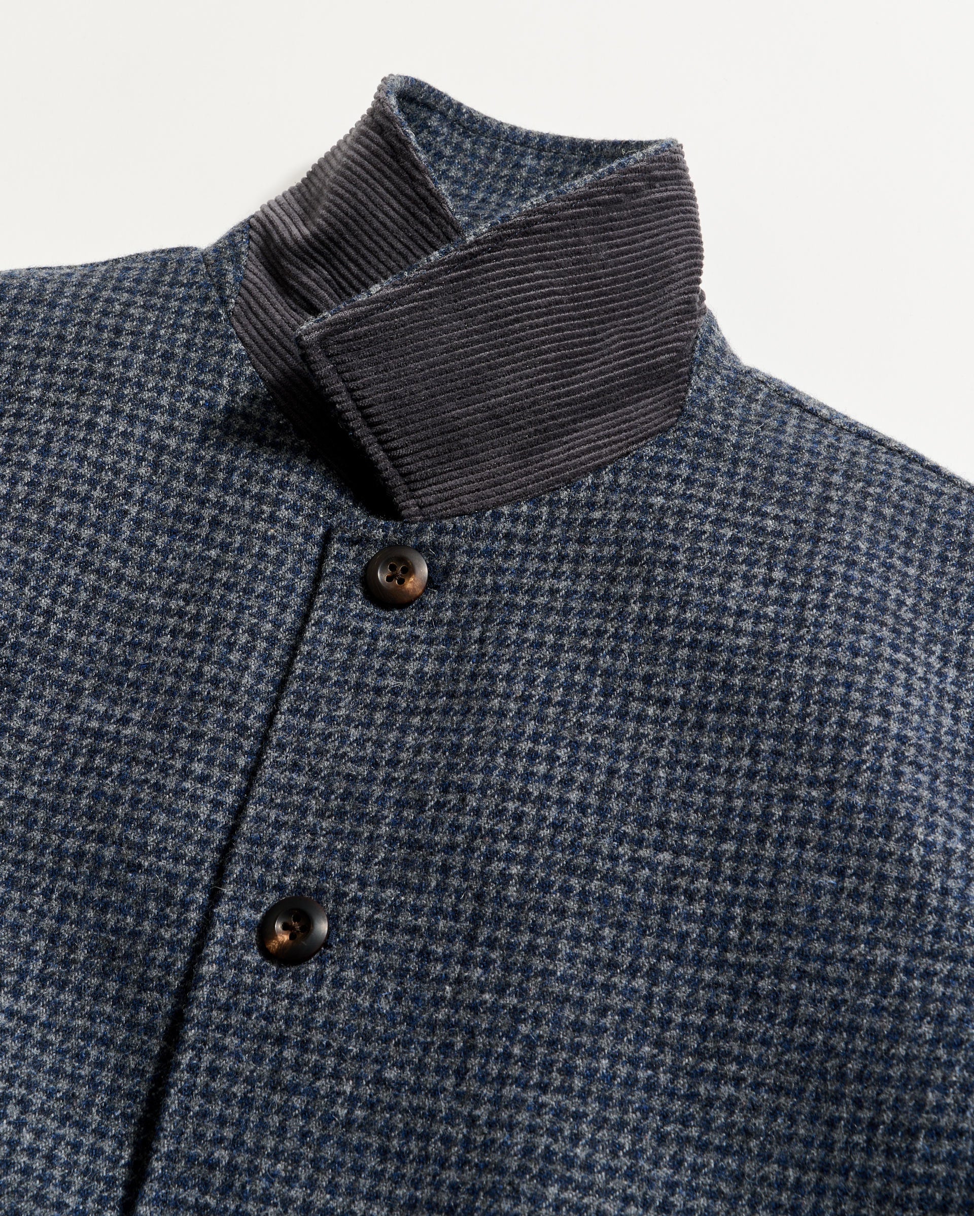 Close-up of a blue and grey houndstooth patterned coat with brown buttons and a contrasting dark gray corduroy collar, photographed against a plain white background.