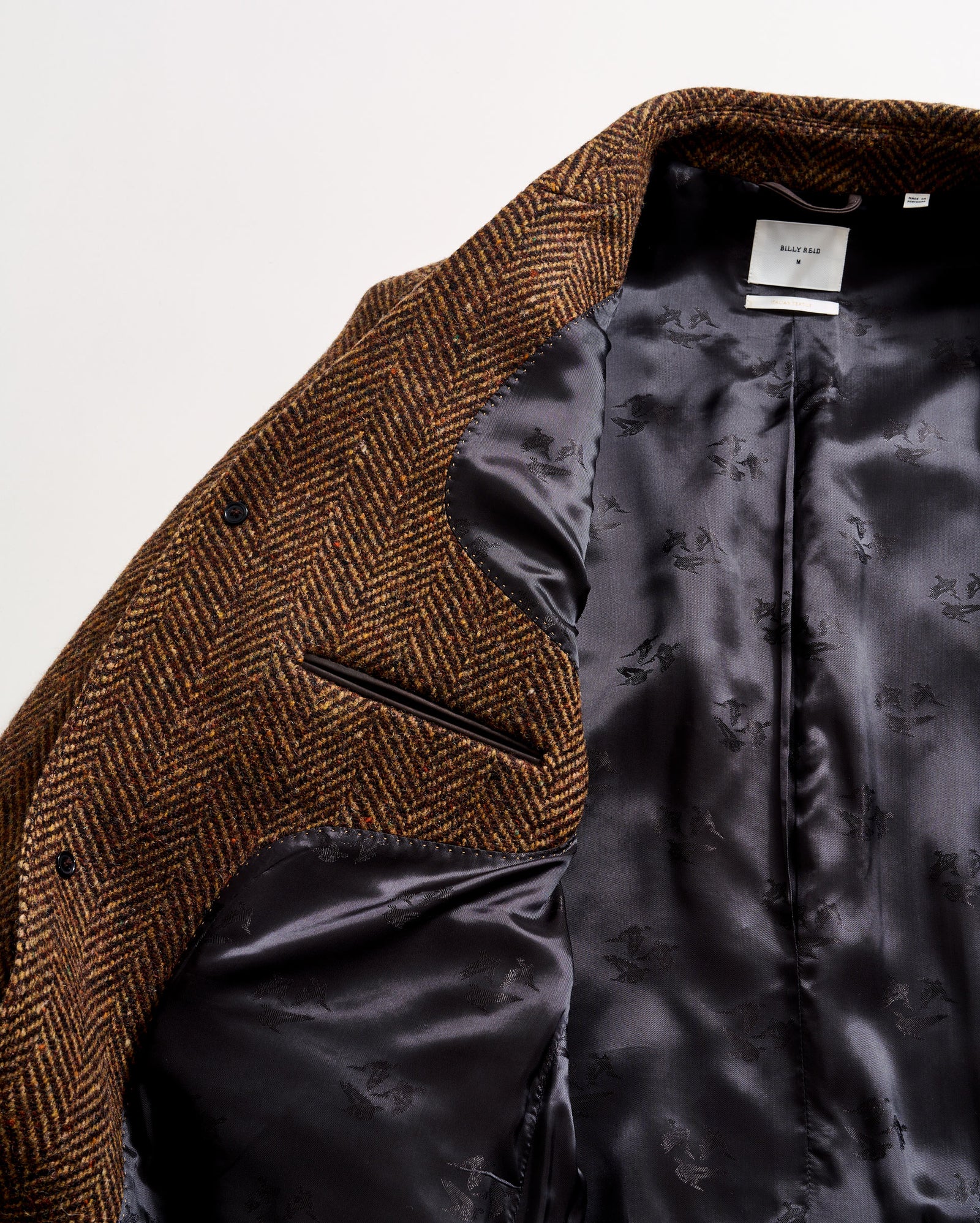 A close-up of a brown herringbone-patterned blazer, partially open to reveal a smooth, dark gray satin lining with subtle floral designs and a white label at the collar.