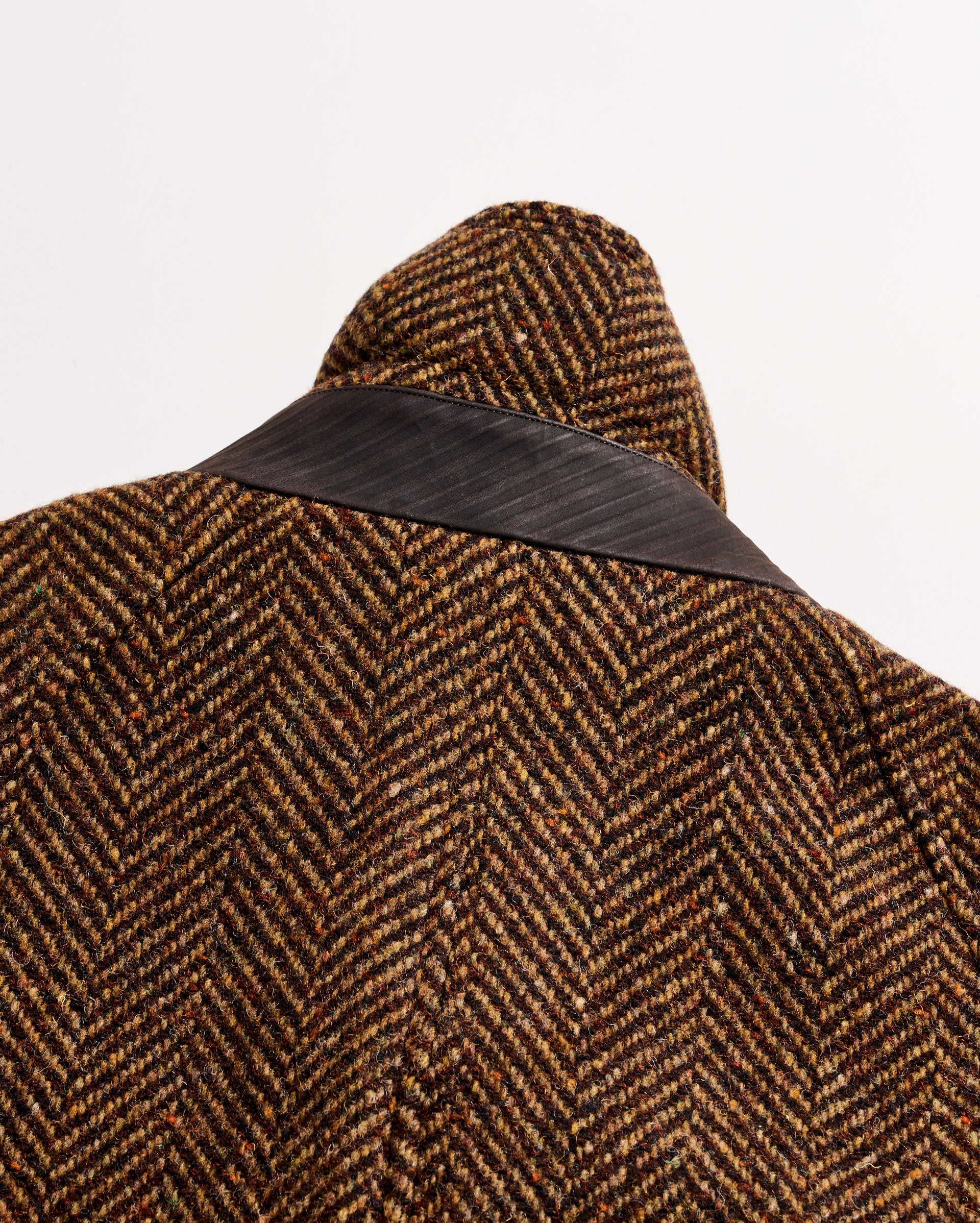 Close-up of the back of a brown and black herringbone-patterned coat with a dark, smooth leather collar on a white background.