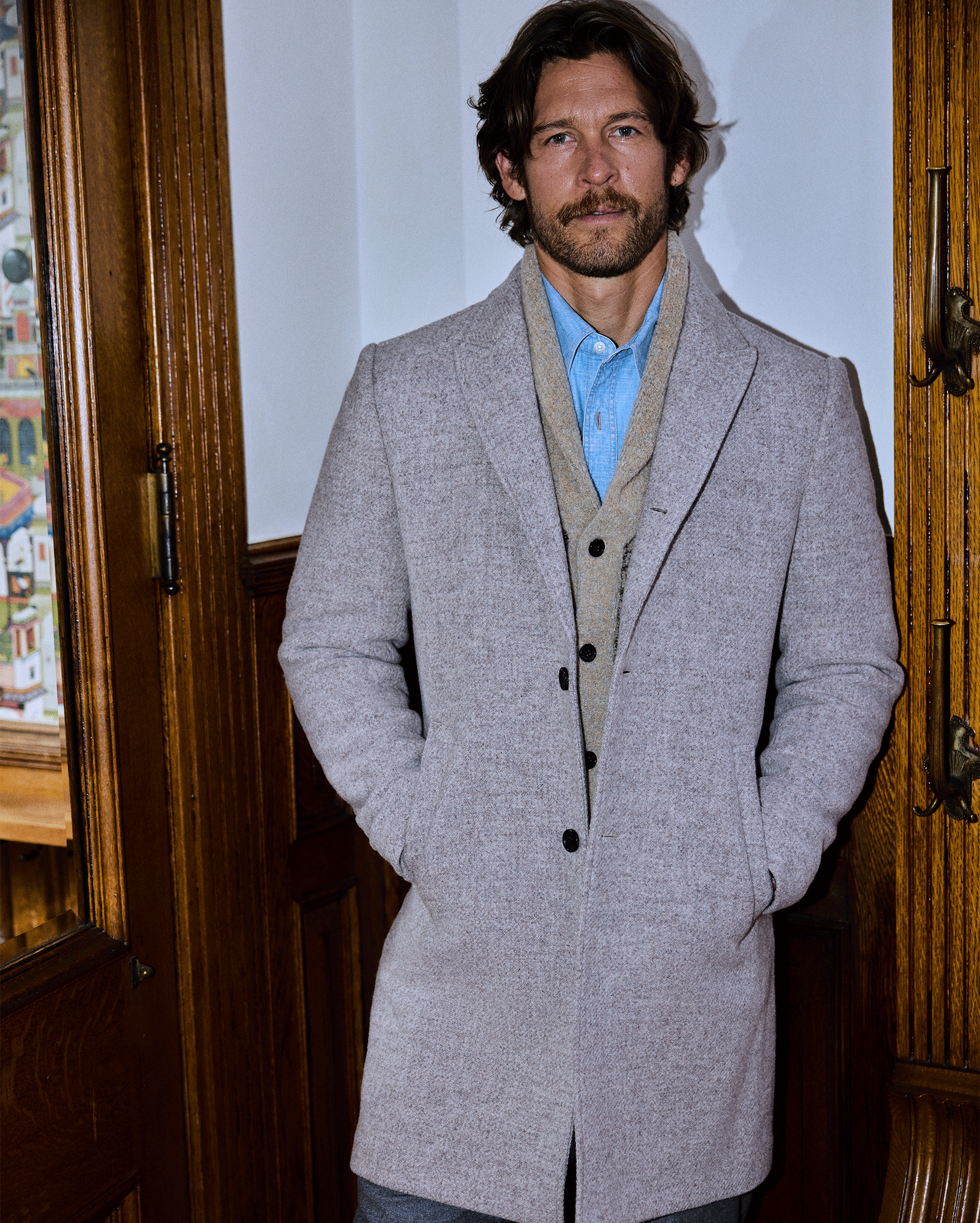 A man with brown hair and a beard stands indoors, wearing a light gray overcoat, a beige cardigan, and a blue shirt. He has his hands in his coat pockets and is positioned near wooden doors.