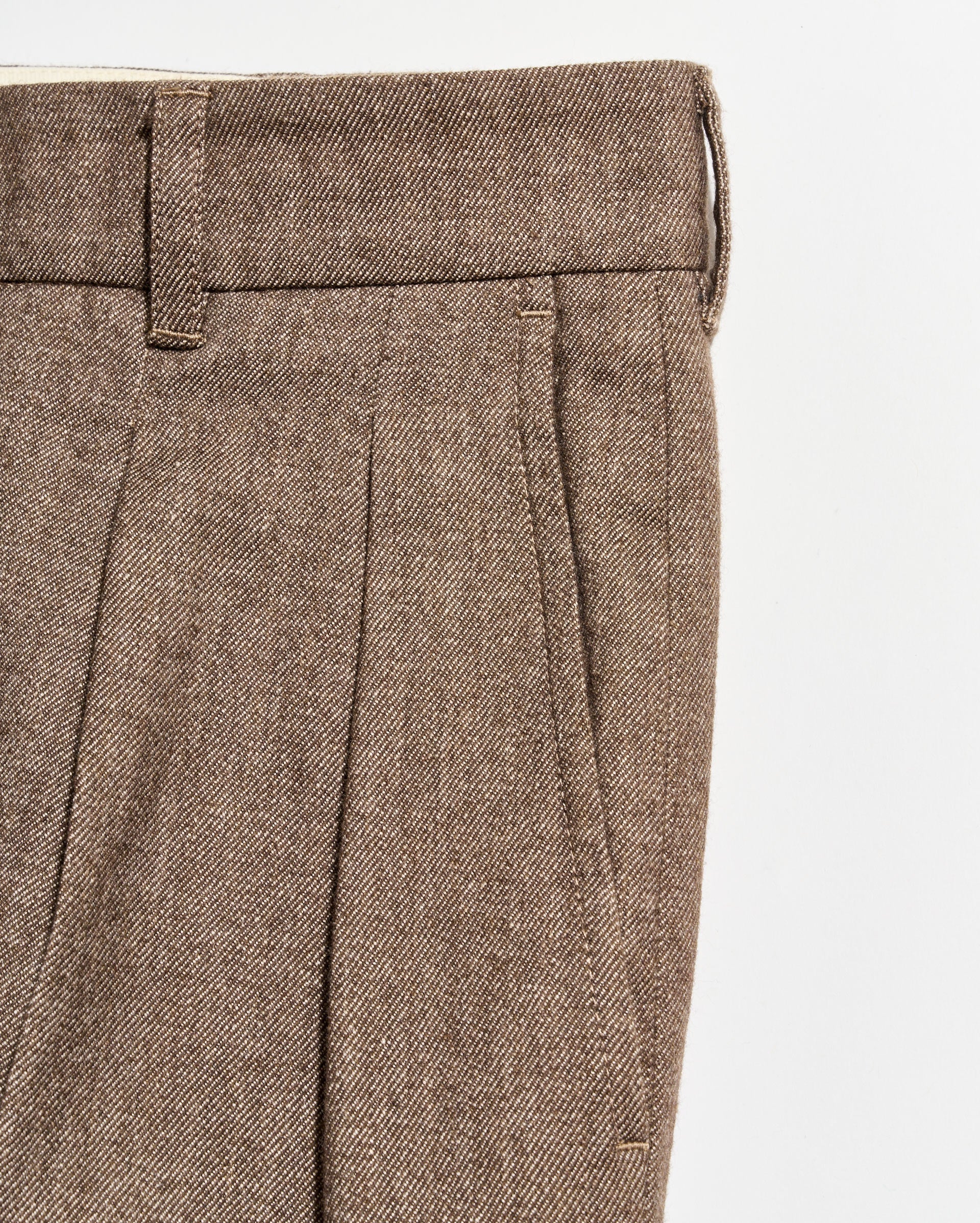 Close-up of brown tweed trousers with pleats, a waistband with belt loops, and a side pocket visible against a white background.