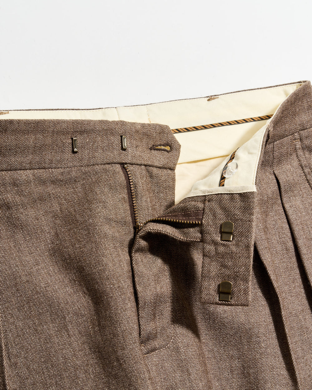 Close-up of brown pleated pants with a partially unzipped fly, showing the inner cream-colored waistband, two metal hook fasteners, and a zipper.