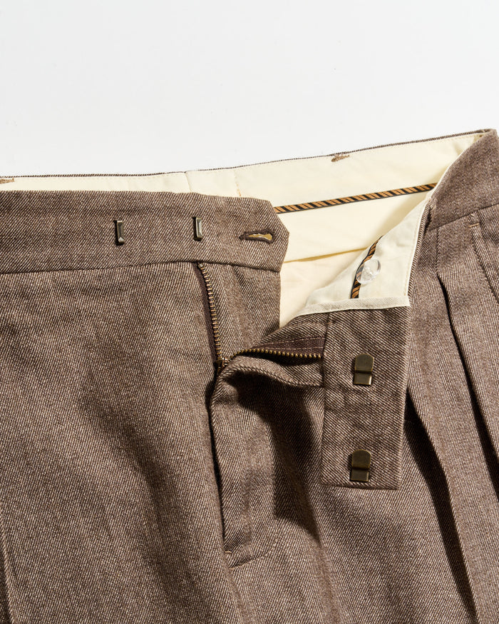 Close-up of brown pleated pants with a partially unzipped fly, showing the inner cream-colored waistband, two metal hook fasteners, and a zipper.