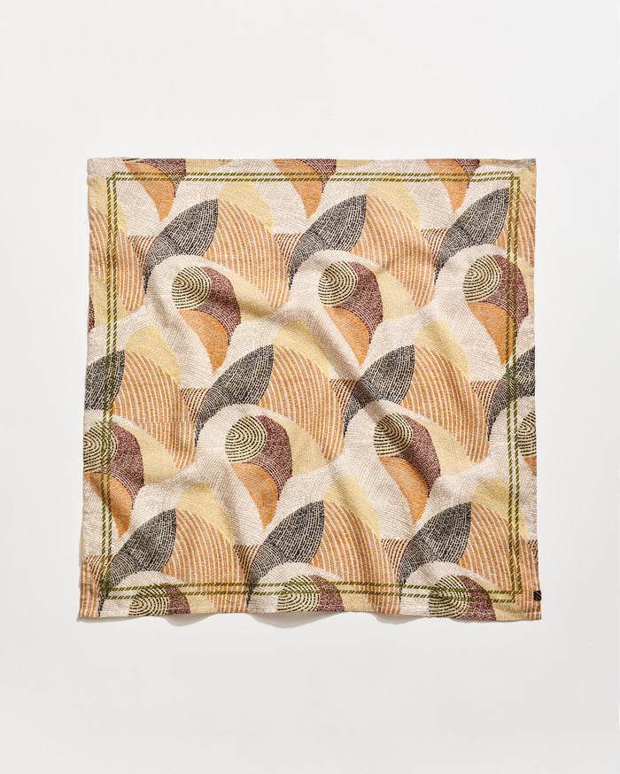 A square cloth napkin with an abstract, geometric pattern in earthy tones of beige, brown, rust, and olive green, featuring curved lines and shapes on a white background. The napkin is slightly wrinkled.