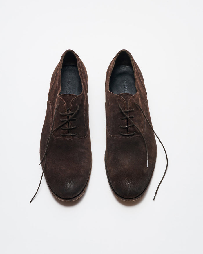 A pair of brown suede lace-up dress shoes is displayed on a white background, with the left shoe fully laced and the right shoes laces partially undone.