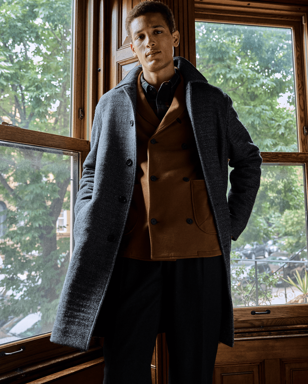 A man stands indoors in front of large wooden-framed windows, wearing a grey overcoat over a brown blazer and black pants. Green trees and buildings are visible outside.