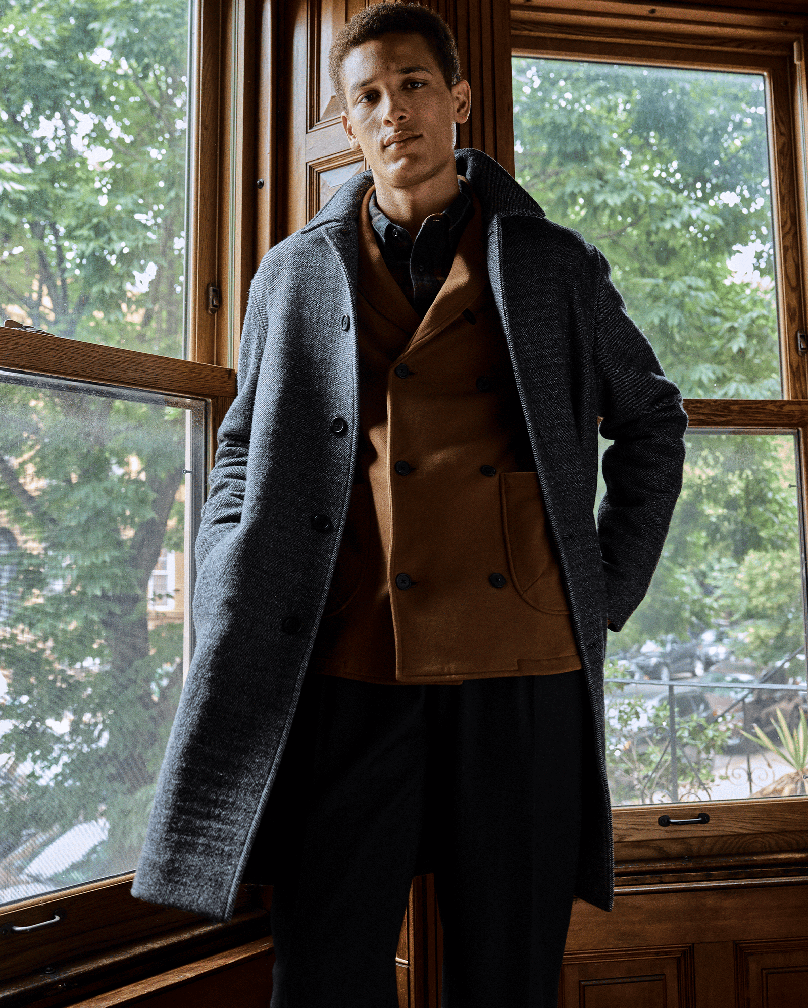 A man stands indoors in front of large wooden-framed windows, wearing a grey overcoat over a brown blazer and black pants. Green trees and buildings are visible outside.