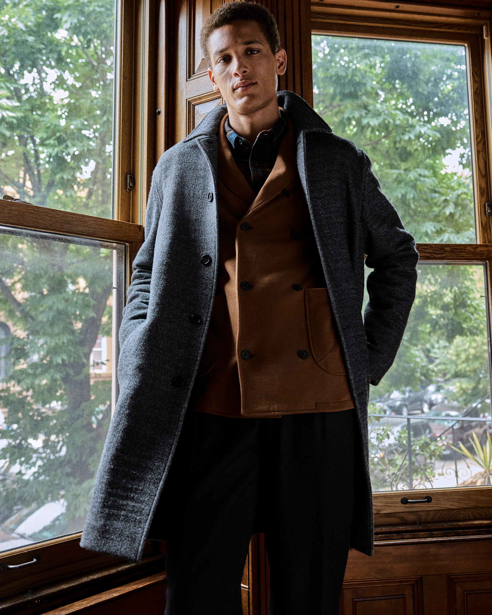 A man stands indoors in front of large wooden-framed windows, wearing a grey overcoat over a brown blazer and black pants. Green trees and buildings are visible outside.