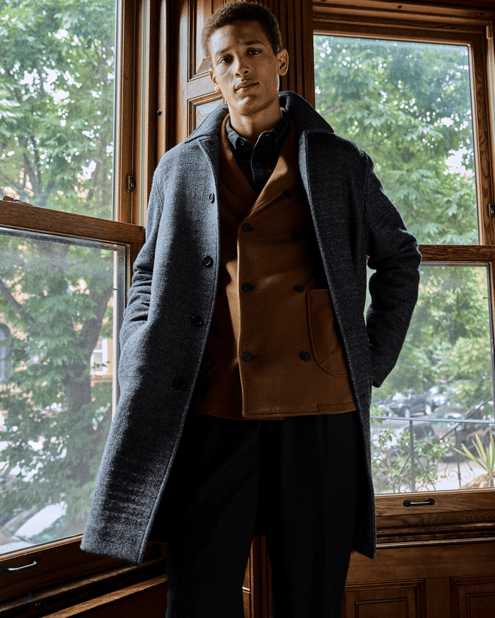 A man stands indoors in front of large wooden-framed windows, wearing a grey overcoat over a brown blazer and black pants. Green trees and buildings are visible outside.