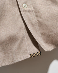 Close-up of a light beige cotton button-down shirt showing the fabric texture, a cream-colored button, and a striped brand label at the hem.