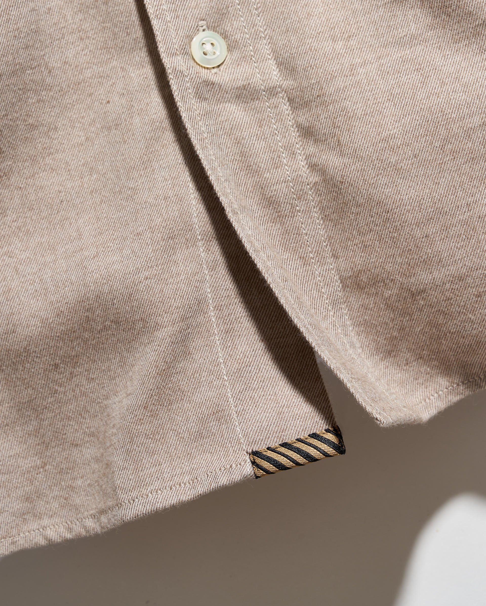 Close-up of a light beige cotton button-down shirt showing the fabric texture, a cream-colored button, and a striped brand label at the hem.