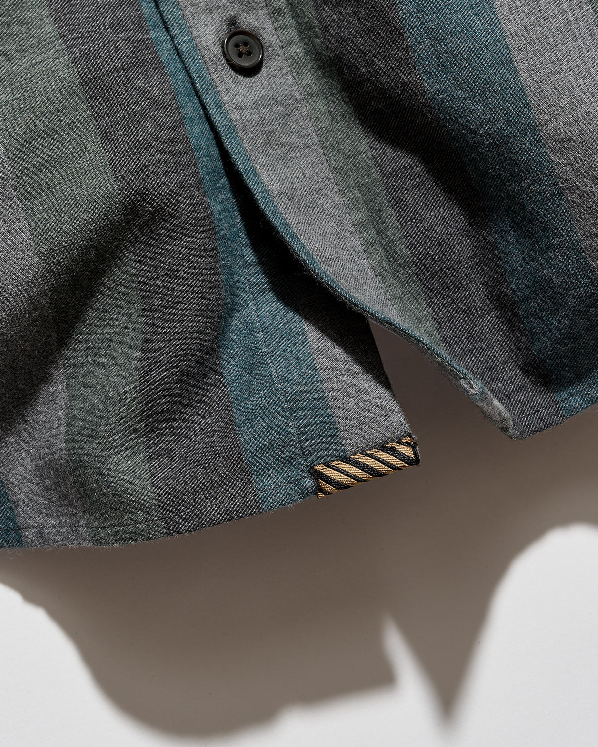 Close-up of a striped button-up shirt in muted blue-gray, charcoal, and slate tones, with a visible striped inner brand label near the hem.