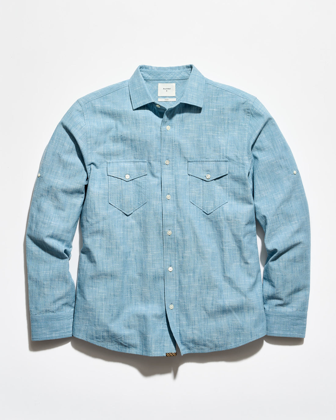 A light blue chambray long-sleeve button-up shirt with two chest flap pockets, laid flat against a white background.