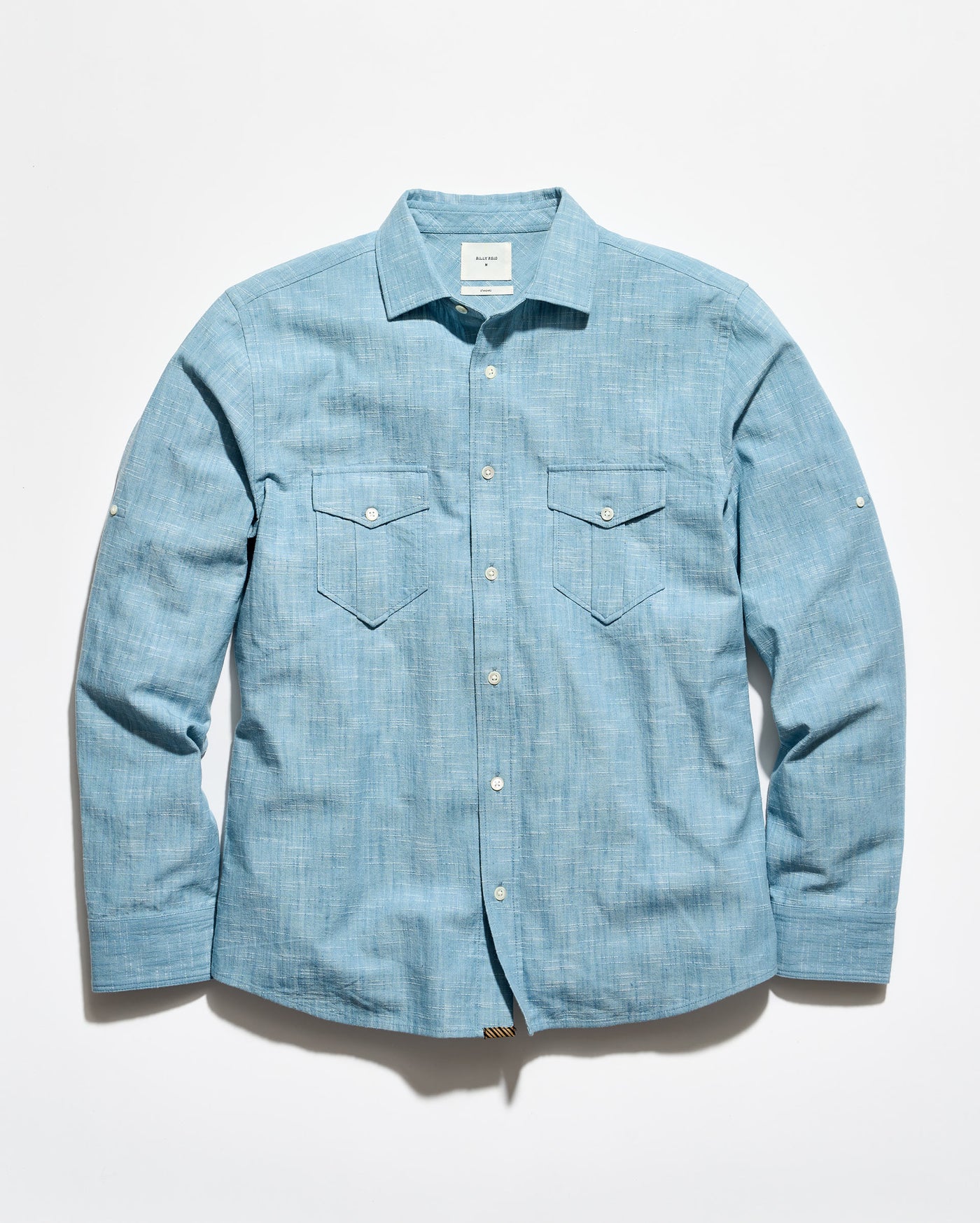 A light blue chambray long-sleeve button-up shirt with two chest flap pockets, laid flat against a white background.
