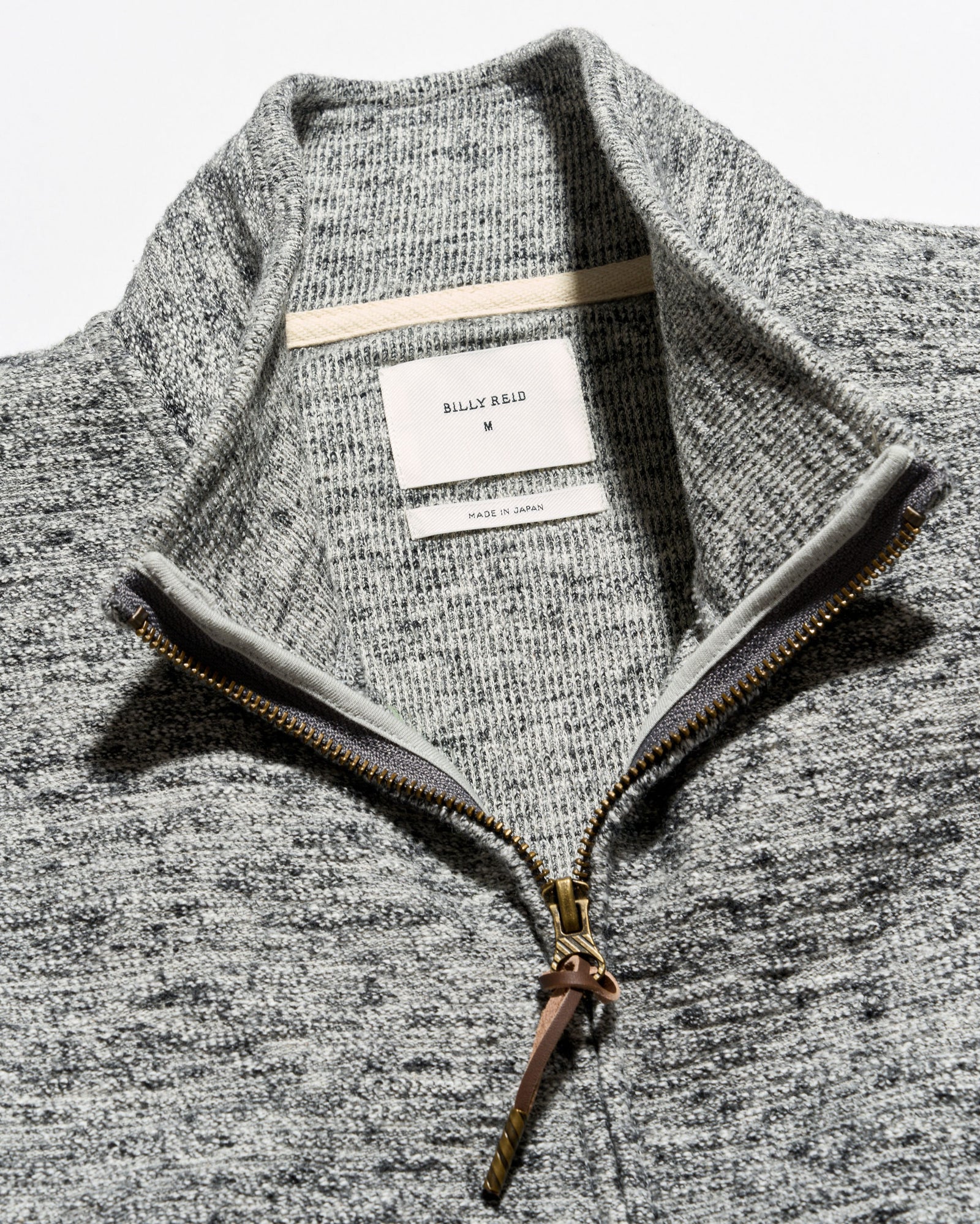 Close-up of a gray heathered knit quarter-zip sweater with a stand collar, showing a white neck label that reads Billie Eilil and a gold-toned zipper.