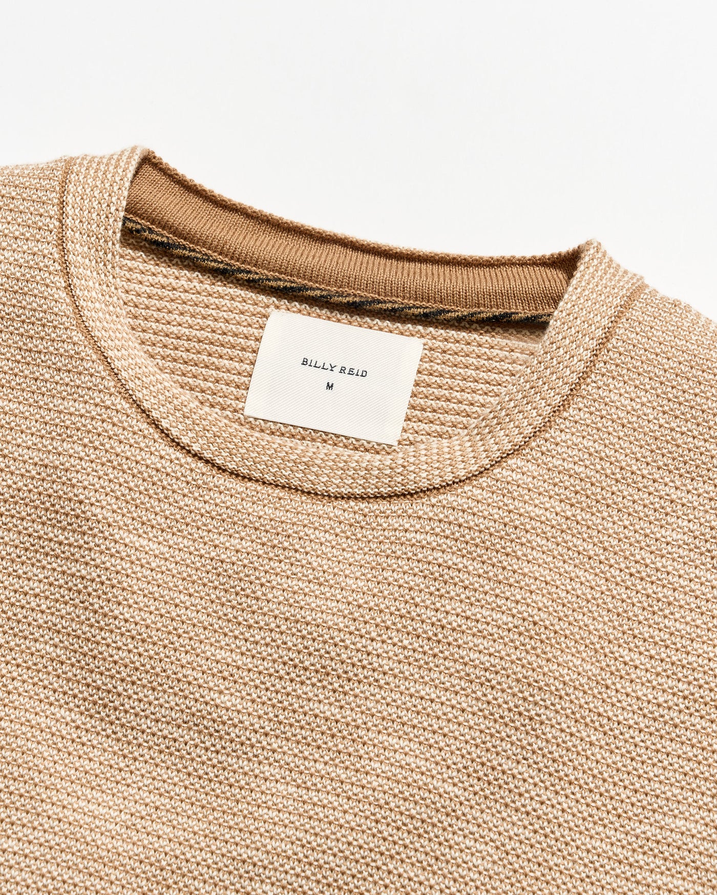 Close-up of a textured knit crewneck sweater in a warm beige/tan color, showing the ribbed collar and a white Billy Reid brand label.