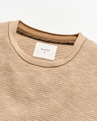 Close-up of a textured knit crewneck sweater in a warm beige/tan color, showing the ribbed collar and a white Billy Reid brand label.