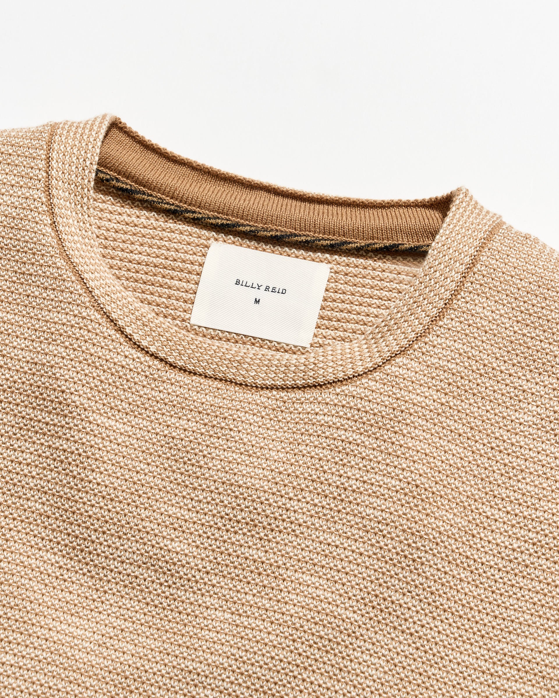 Close-up of a textured knit crewneck sweater in a warm beige/tan color, showing the ribbed collar and a white Billy Reid brand label.