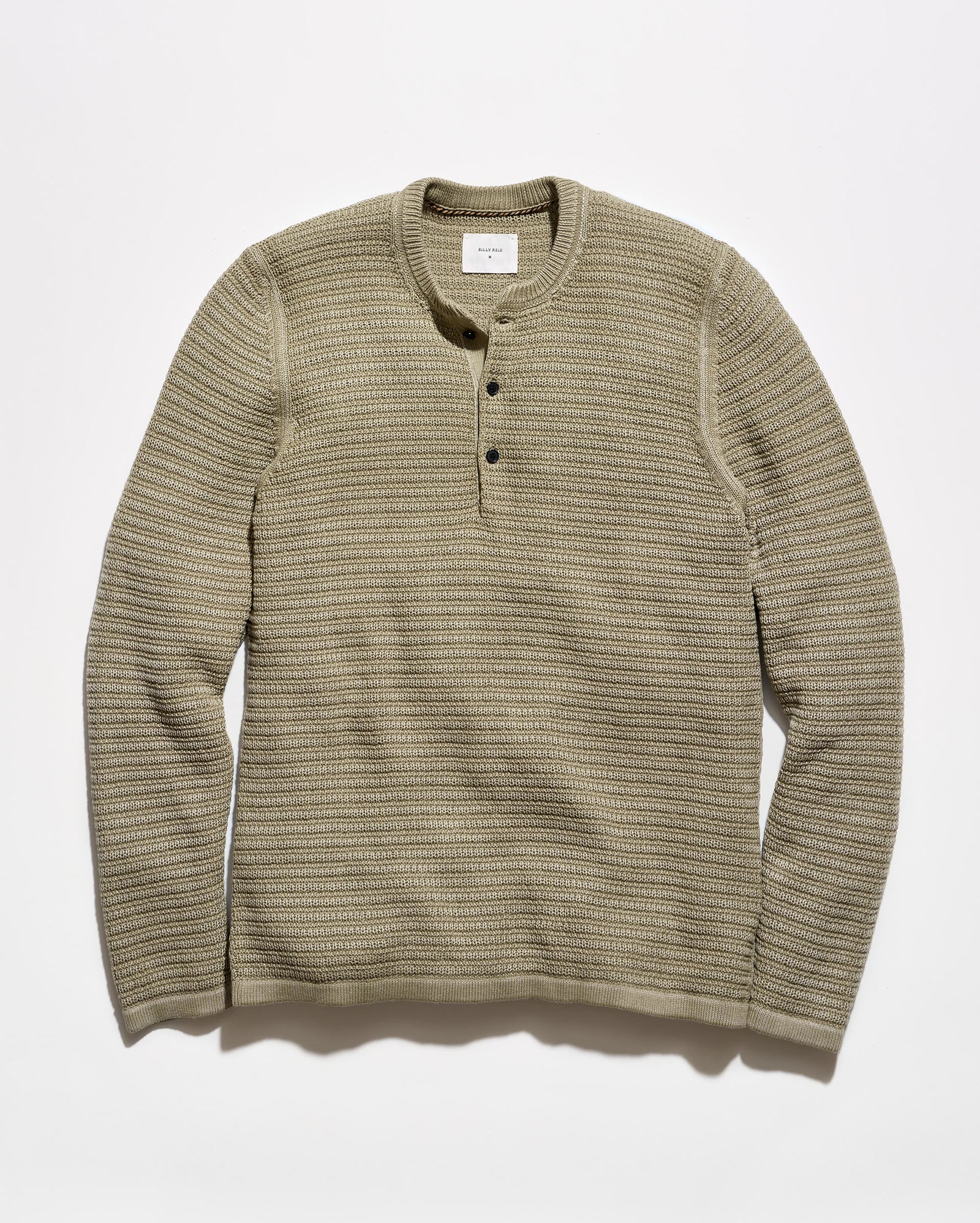 A ribbed knit long-sleeve henley shirt in a muted olive-beige color, featuring a three-button placket and a crew neck, laid flat against a light gray background.