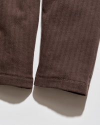 Close-up of the lower legs of dark brown corduroy trousers, showing the ribbed texture and hemstitching against a white background.
