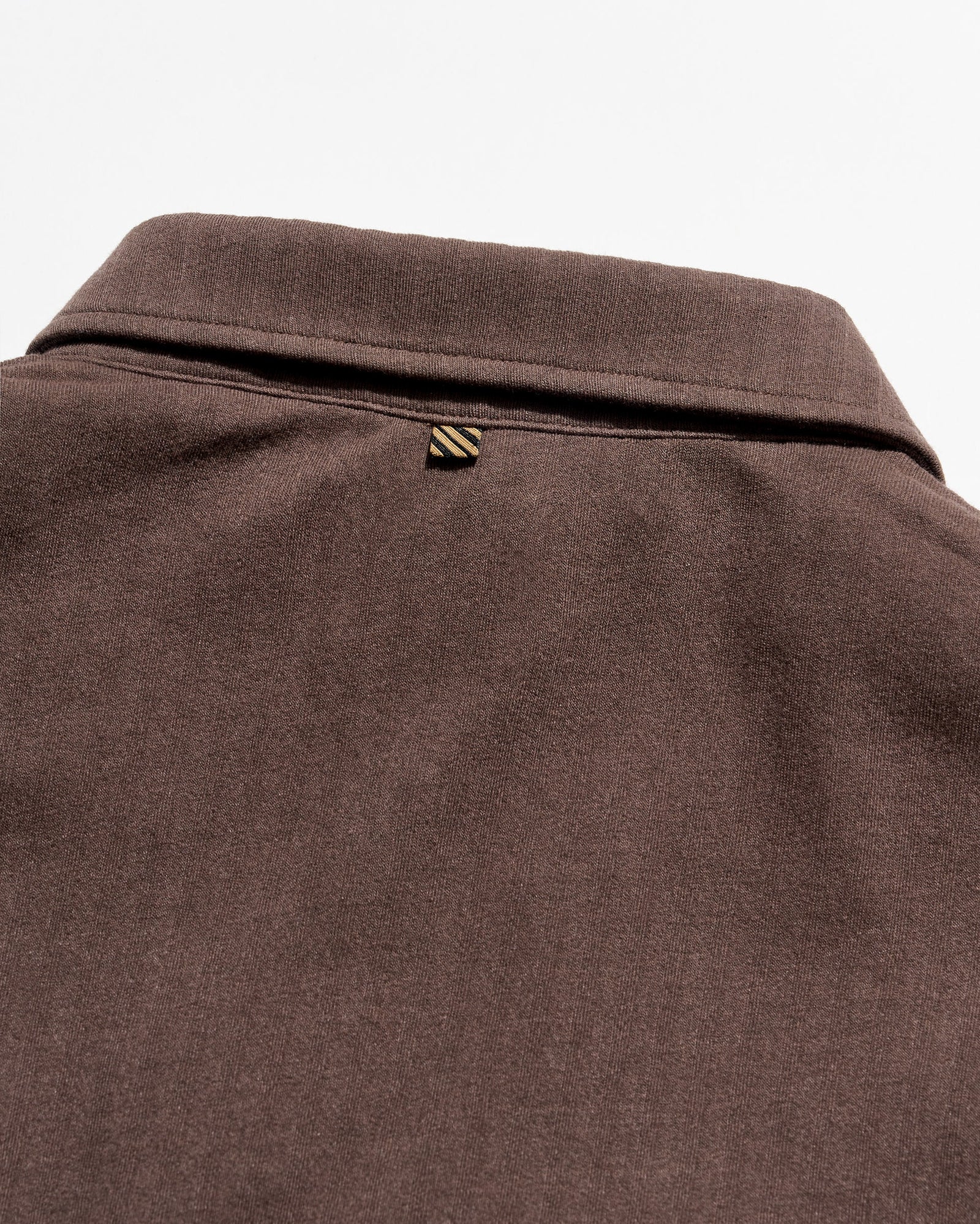 Close-up of a dark brown textured fabric garment, showing the back of a collared shirt or sweater with a small striped triangular brand tab near the collar.