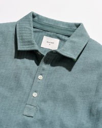 Close-up of a slate blue-green polo shirt by Billy Reid, showing the textured fabric, collar, and white buttons.