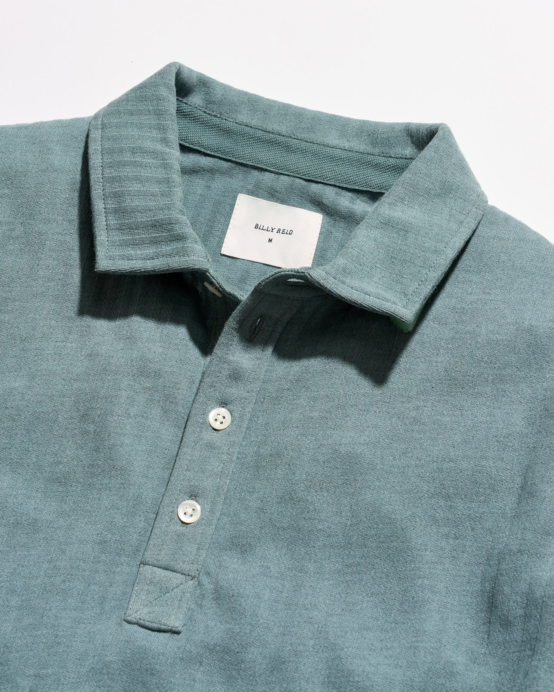 Close-up of a slate blue-green polo shirt by Billy Reid, showing the textured fabric, collar, and white buttons.